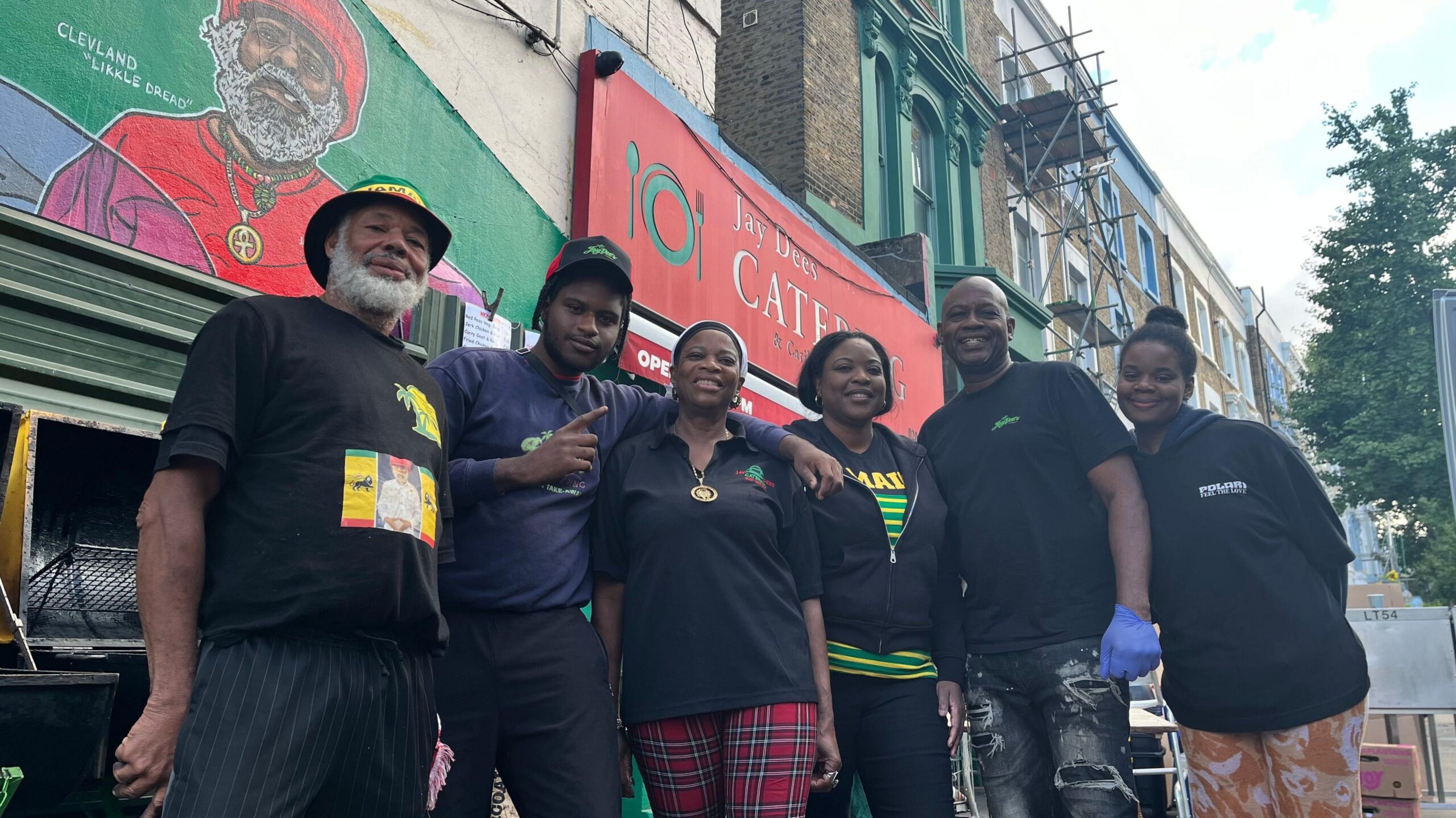 A group of three men and three women stand in a row with their arms around each other in front of Jay Dee's Catering.