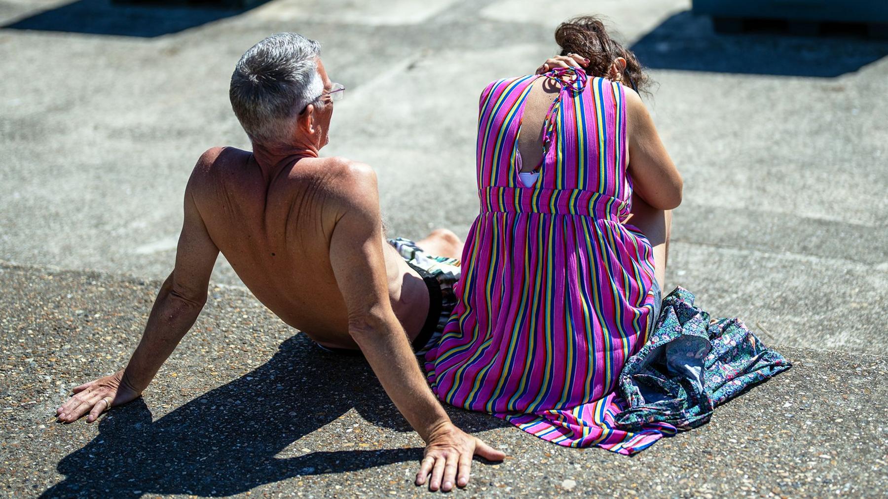 A man and a woman sit in the sun. The man is topless and leans back, the woman is in a purple sun dress has her head lowered and shields herself from the sun