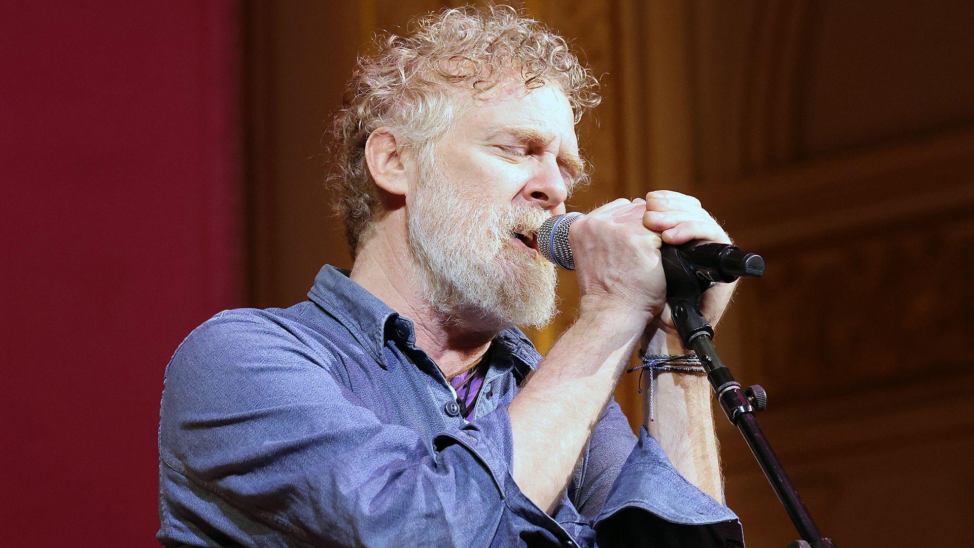 Glen Hansard has short curly reddish/brown/grey hair and a greyr beard and is singing with two hands on the microphone and his eyes closed