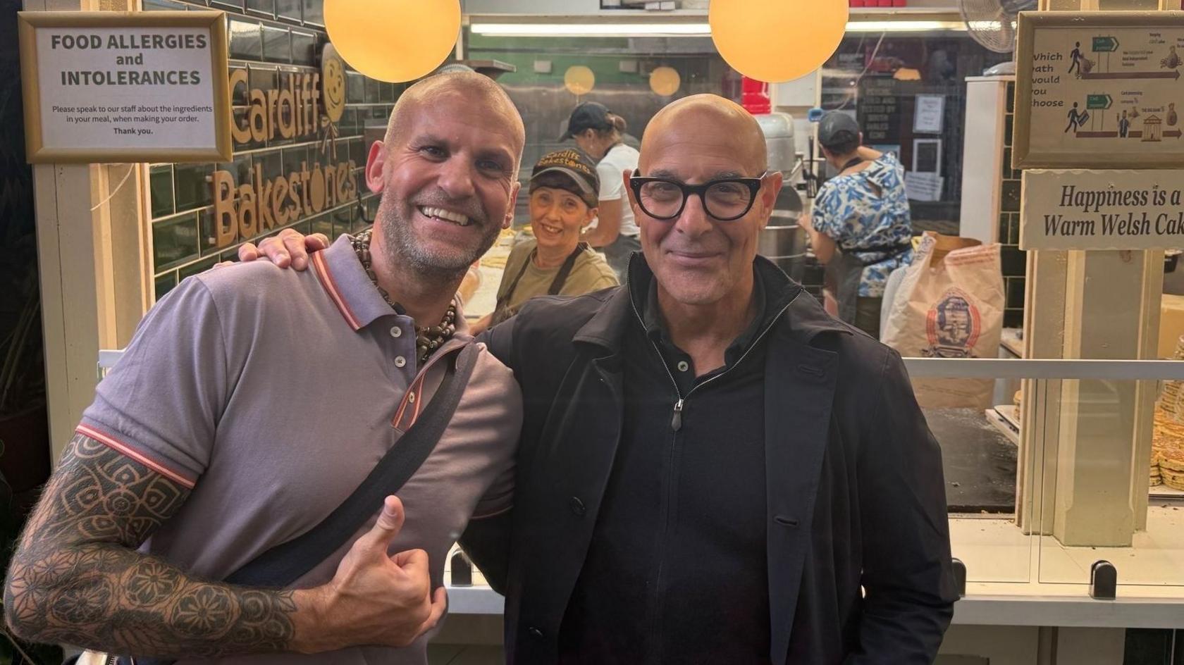 Owner of the Welsh cake store stood next to actor Stanley Tucci. He has a full sleeve tattoo and a bag strap across his body. Tucci is wearing a black jacket over a black quarter zip jumper. There is a staff member smiling in the background with a stack of Welsh cakes on the right. They sit under as sign reading: Happiness is a Warm Welsh Cake