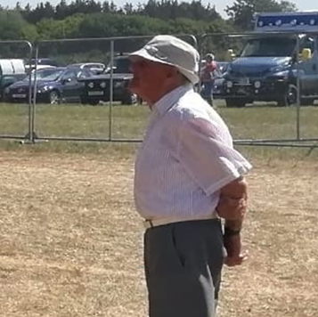 An old man wearring suit trousers and shirt, and a bucket hat, standing side on in a field.