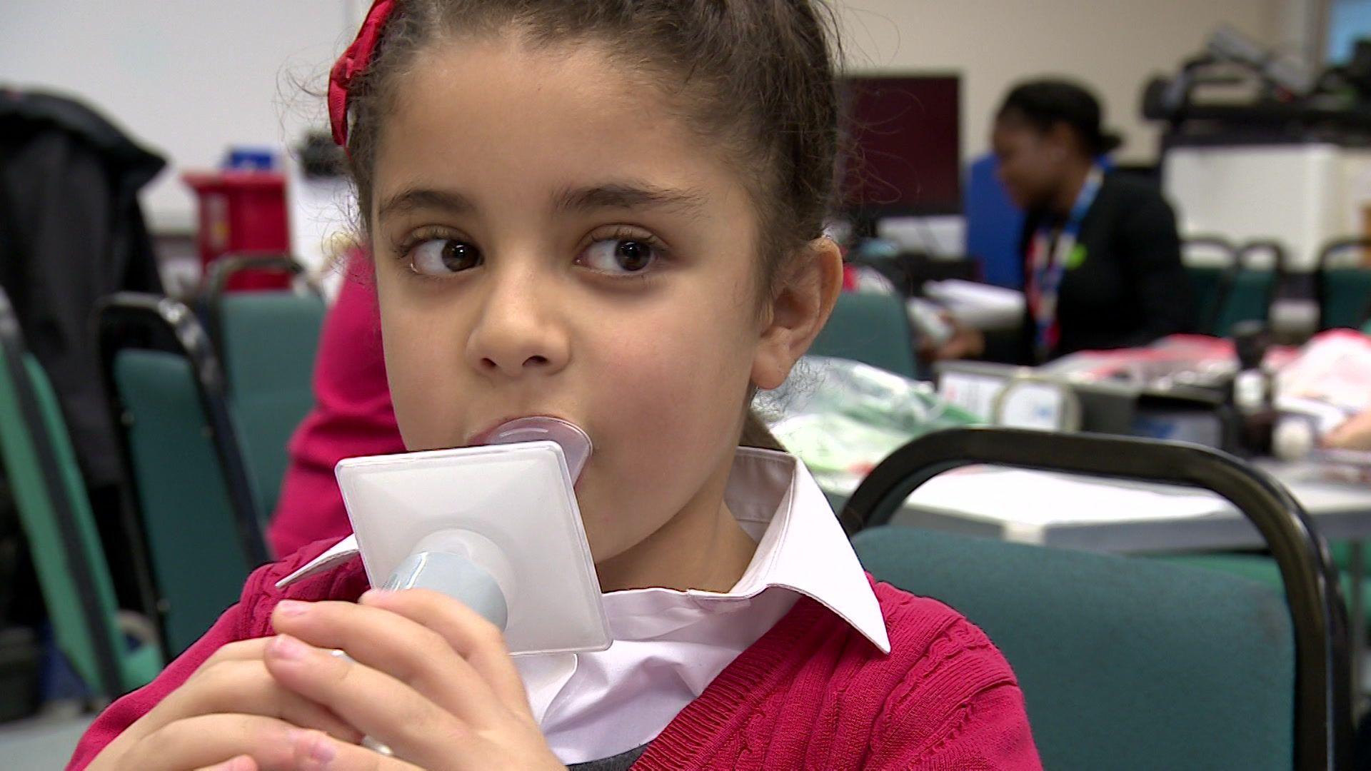Young child with brown hair, blowing air into plastic measuring device. She wears a red ribbon and red cardigan and a white shirt, She is in school, sitting on a green chair.