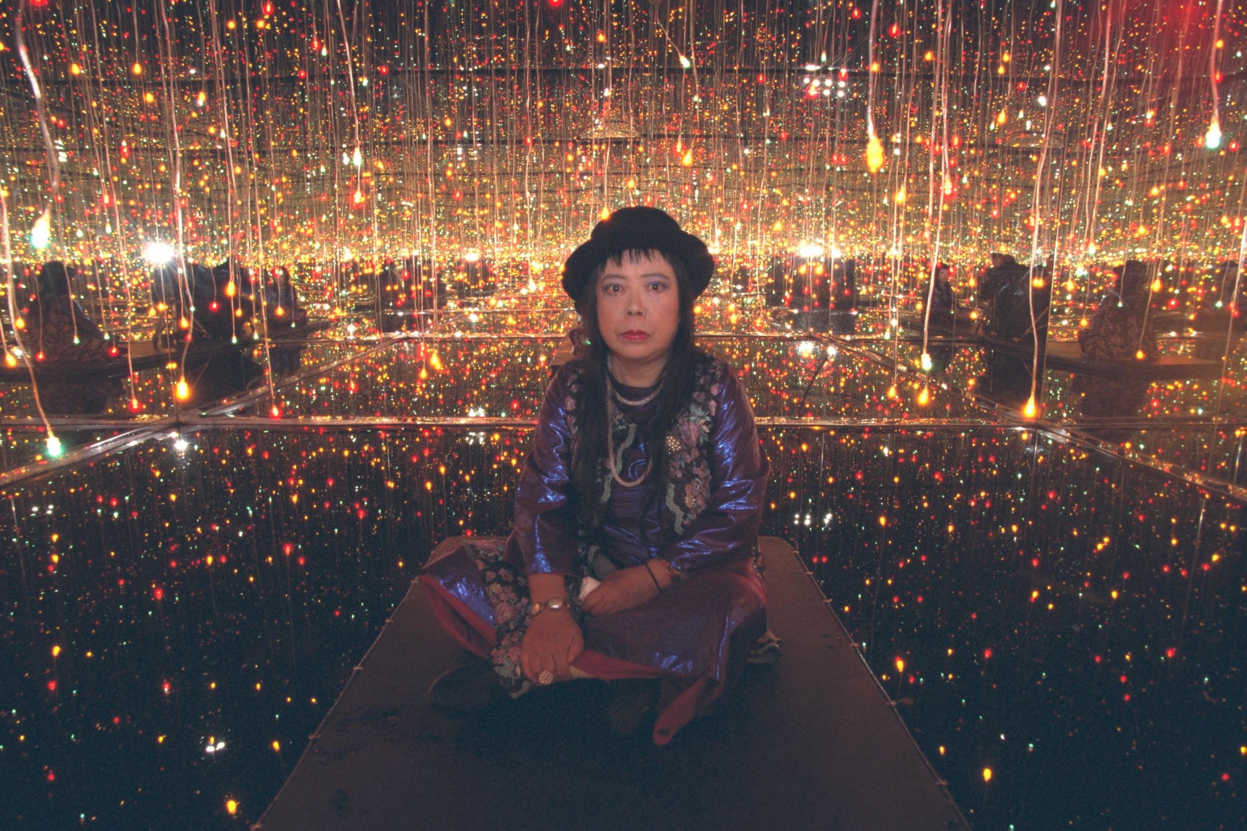 Yayoi Kusama in 2001 seated on the floor of one of her installation rooms, surrounded by hundreds of small coloured lights and mirrors