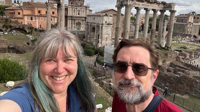 A selfie image showing Lynda and Joe Rousseau in front of ancient Roman ruins and columns. Lynda is smiling and has grey hair with bangs and teal streaks. Joe is wearing black sunglasses and has a grey and black beard, and short brown hair.