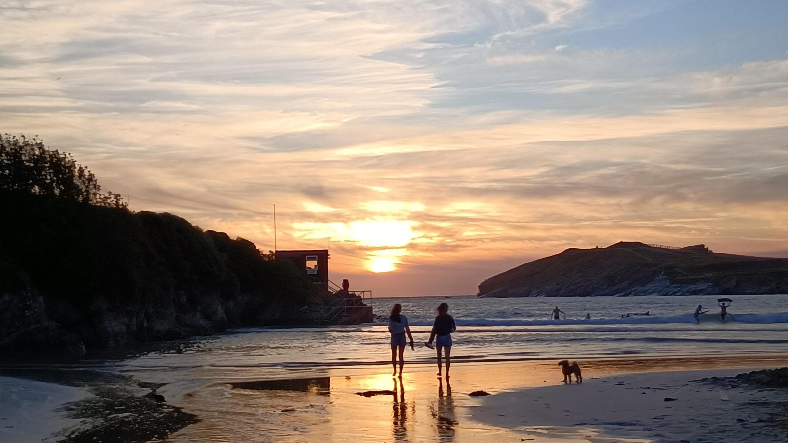 Two people and a dog stroll along the shoreline as the sun sets over the sea