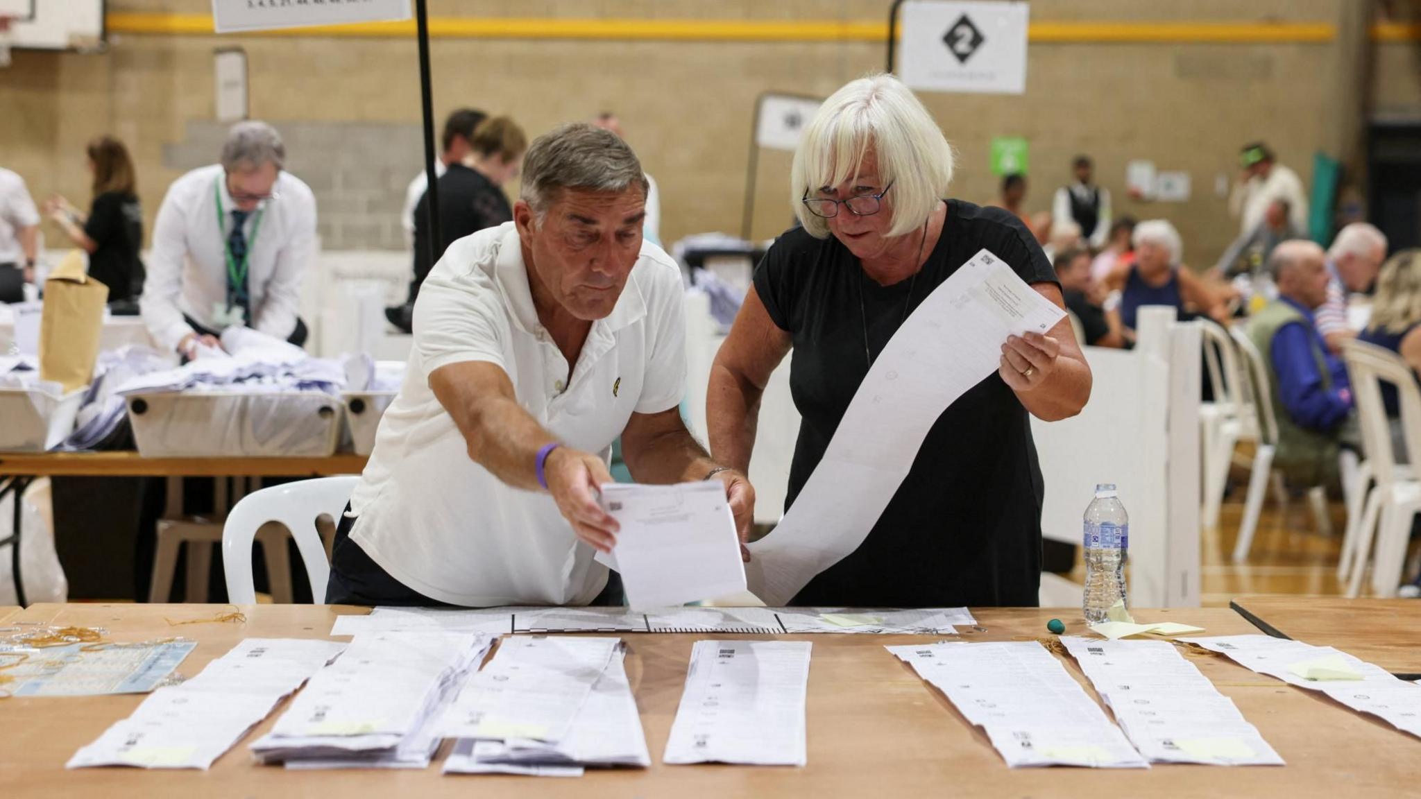 A man and a woman stand behind a trestle table at an election count in a sports hall. They are looking at and holding long ballot papers