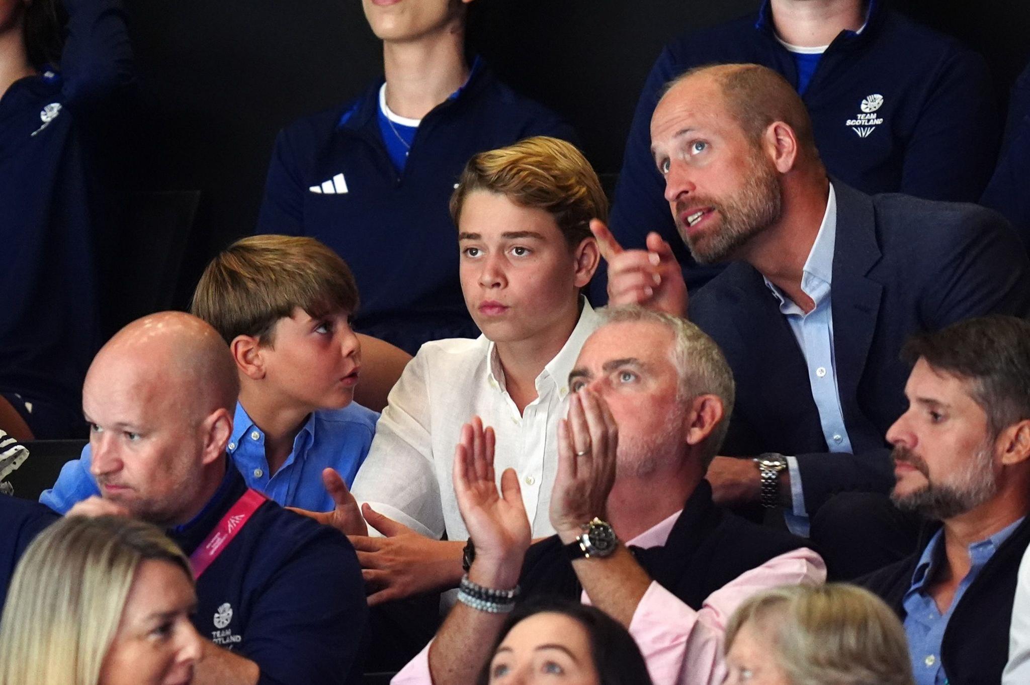 Prince William leans in to speak to his sons George and Louis while the family watch a netball match. William points to something above his eyeline while Louis' attention is on his father. George stares straight ahead.