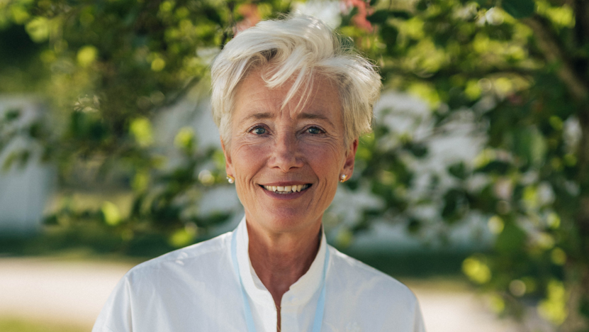 Emma stood outside with a large tree blurred behind her. She wears a white blouse, a light blue medal and has short blonde hair.