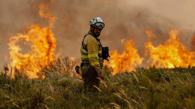 A Spanish firefighter in a field looks back towards the camera. Ahead of him flames flash skywards.