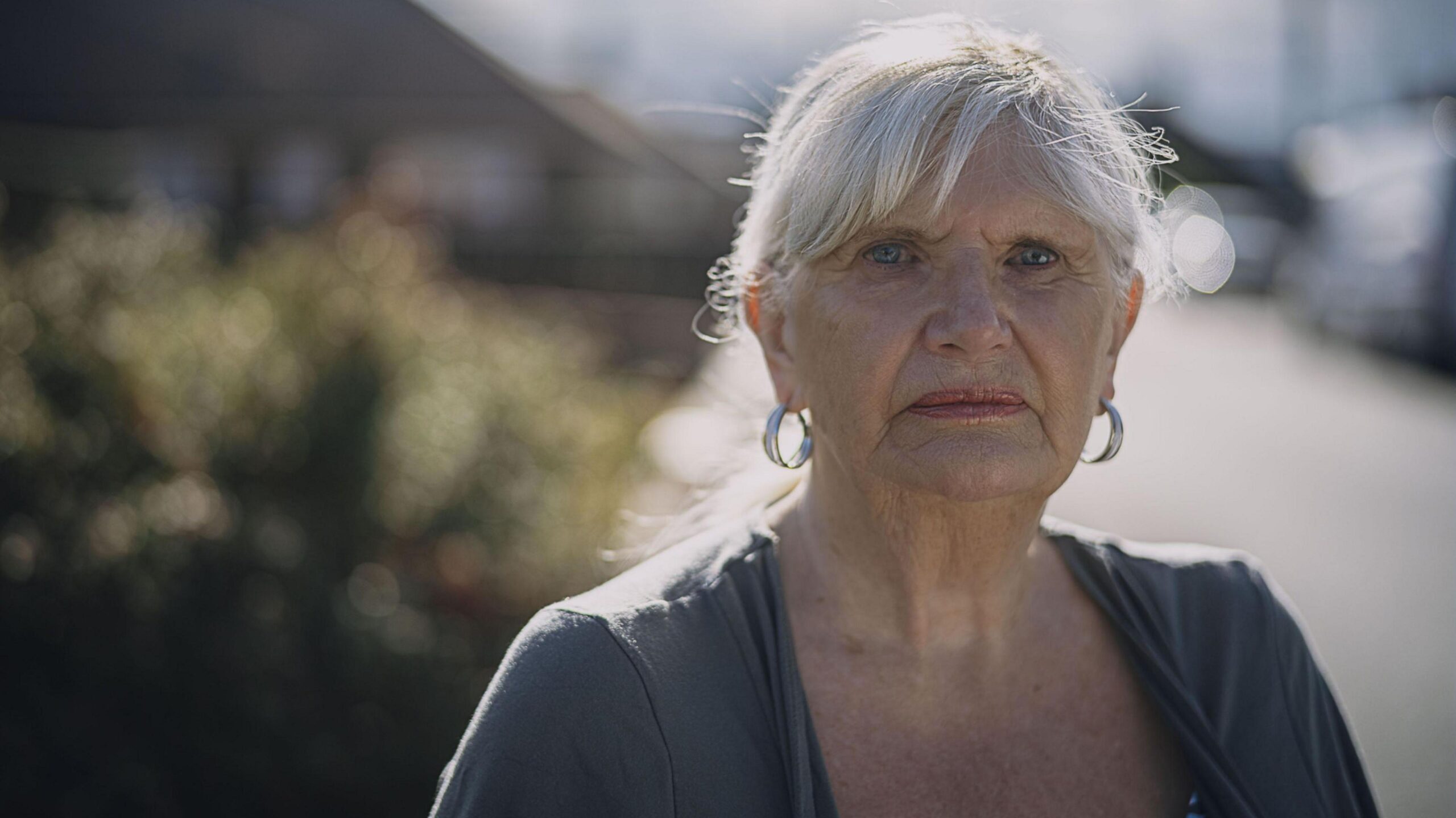 A woman with white hair, tied back in a pony tail. She is looking directly into the camera. She has hooped earrings and is wearing a plain grey top. The background is out of focus.