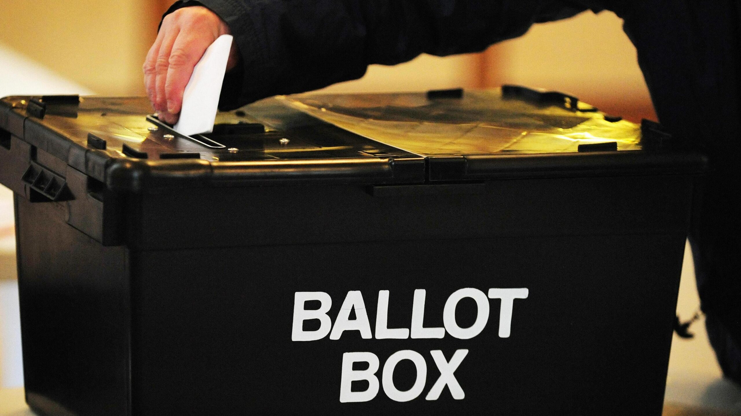 A hand places a ballot paper into a black ballot box.