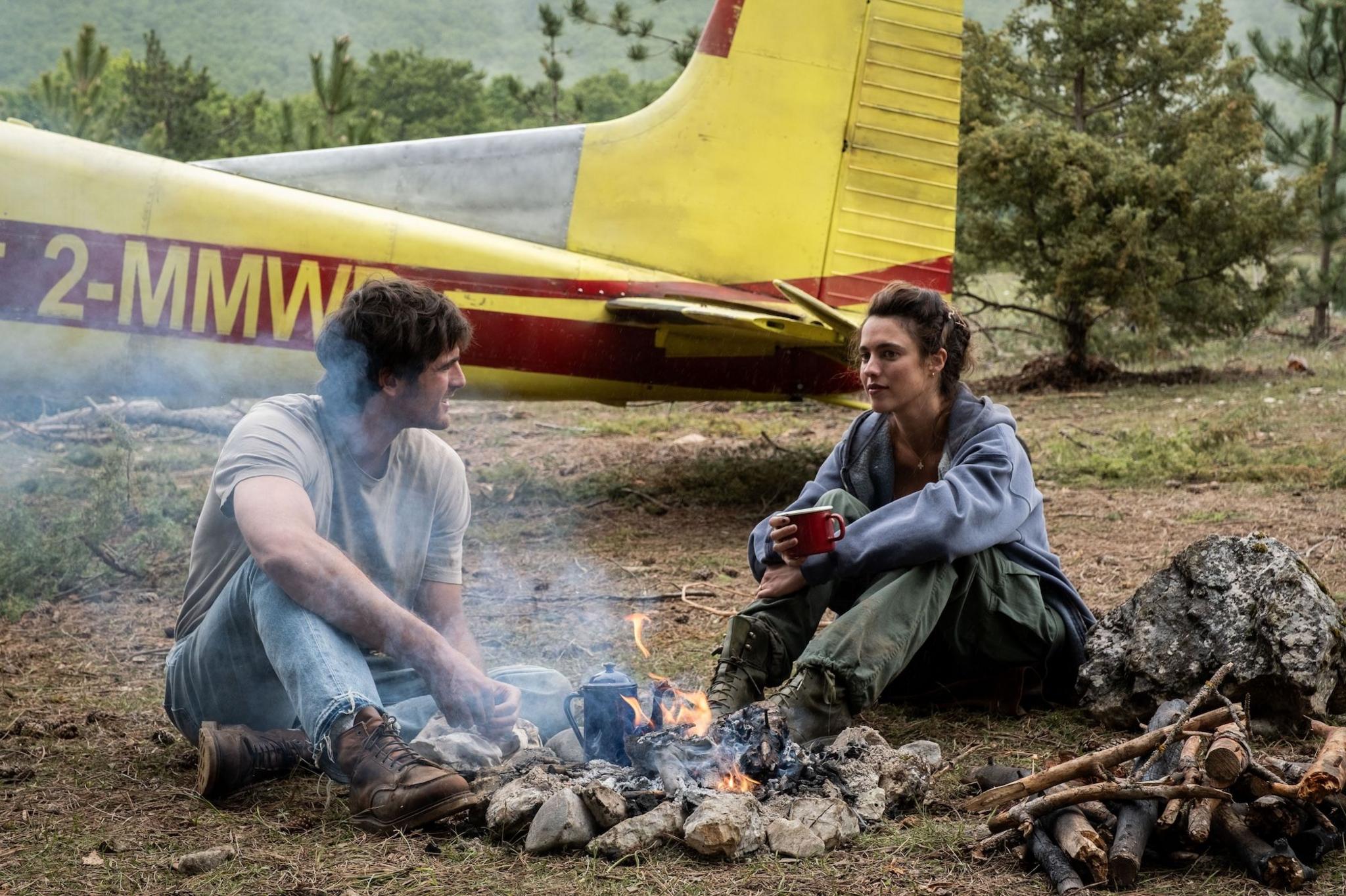 Picture of Jacob Elordi with Margaret Qualley in a still from The Dog Stars. They are sat in front of a fire with a yellow airplane pictured behind them.