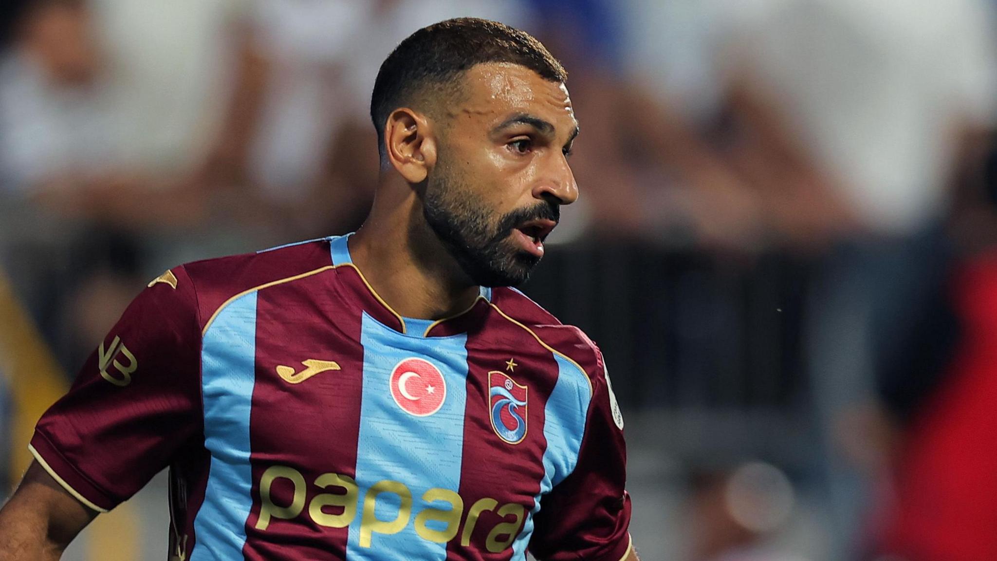 Mohamed Salah, visible from chest up wearing a claret and light blue Trabzonspor shirt with a circular Turkish flag in its centre just under the neck, looks to his left with a focused look during a football match