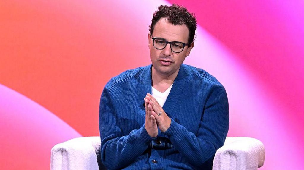 Dario Amodei sitting on-stage at a conference, wearing a blue cardigan and white t-shirt. He is holding his hands clasped in front of him as he speaks.