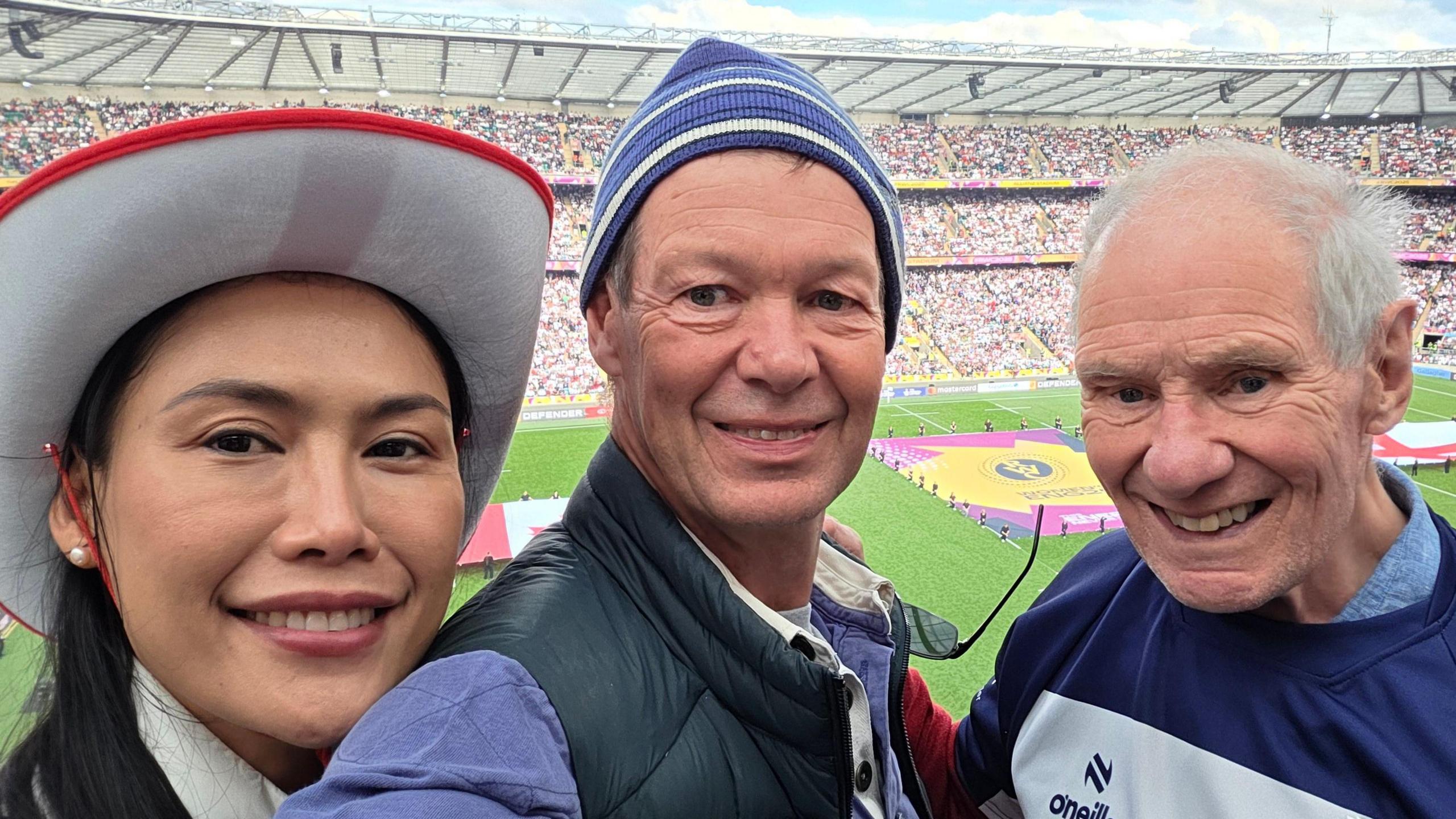 Three people in the foreground pose for a photograph in a large sports stadium. One person wears a red-and-white hat, another wears a striped knitted hat and padded gilet, and a third wears a Bristol Bears rugby shirt. A rugby pitch is visible behind them, with large flags laid out on the grass and stadium seating filled with spectators.