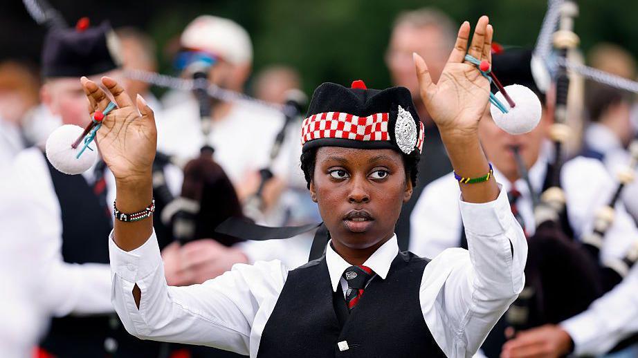 A drum major or mace bearer standing in front of a group of bagpipers. She is wearing a traditional piper outfit with hat