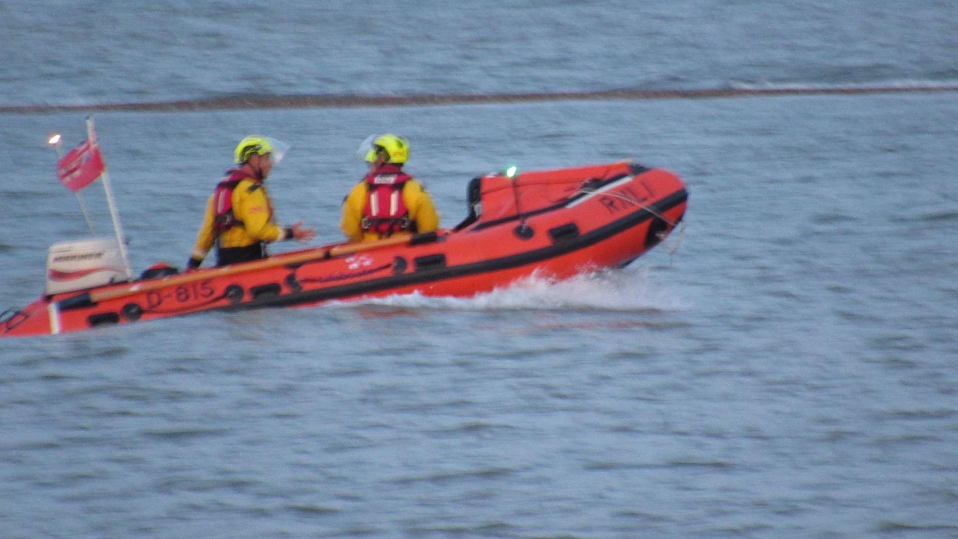 An RNLI boat on the sea at West Mersea. It is orange and has at least two people clad in yellow hi-vis and yellow helmets on board.
