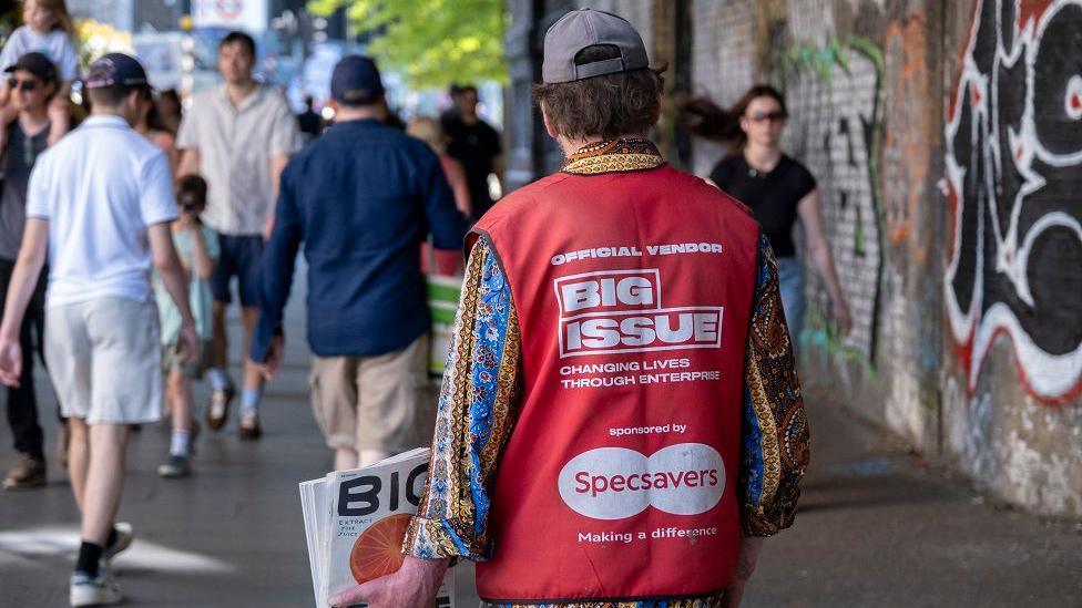 A man, seen from behind, wearing a red Big Issue vest and holding some magazines in his left hand. He is wearing a colourful, patterned shirt underneath and a baseball cap. He is holding up