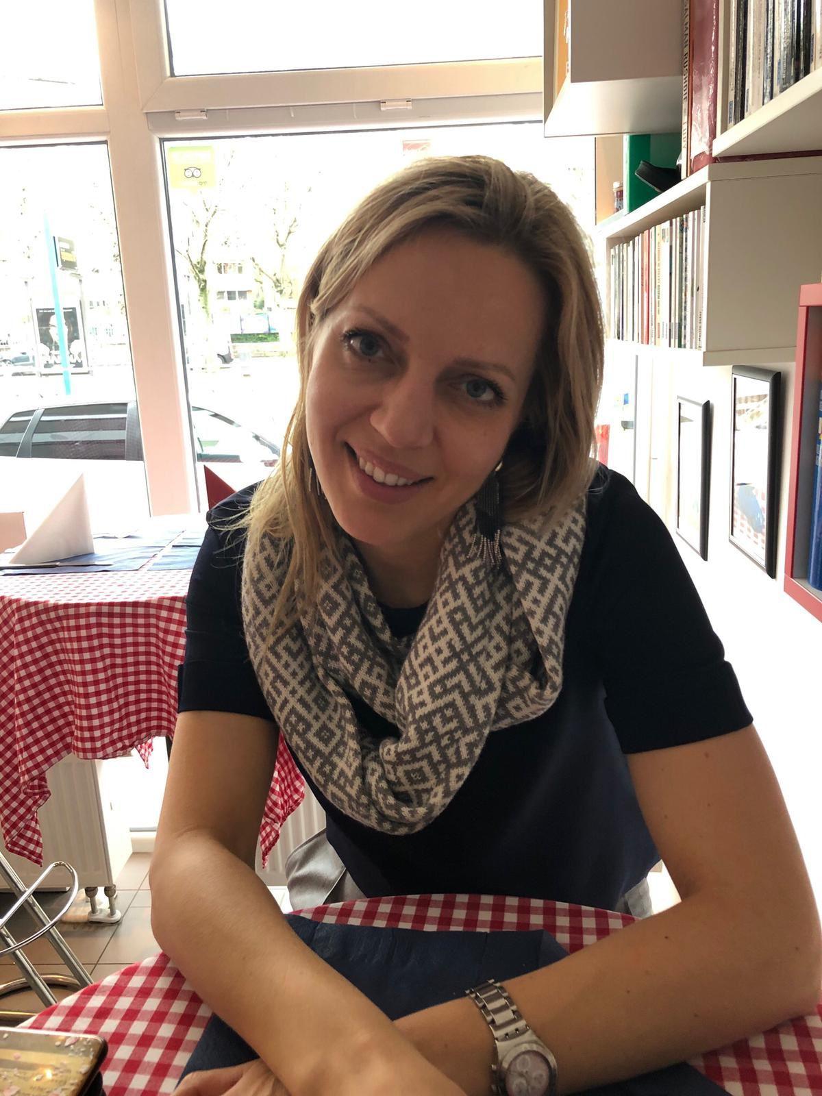 Julija Jakovicka sits at a restaurant table and smiles, she is wearing a black t-shirt and a white and black scarf.