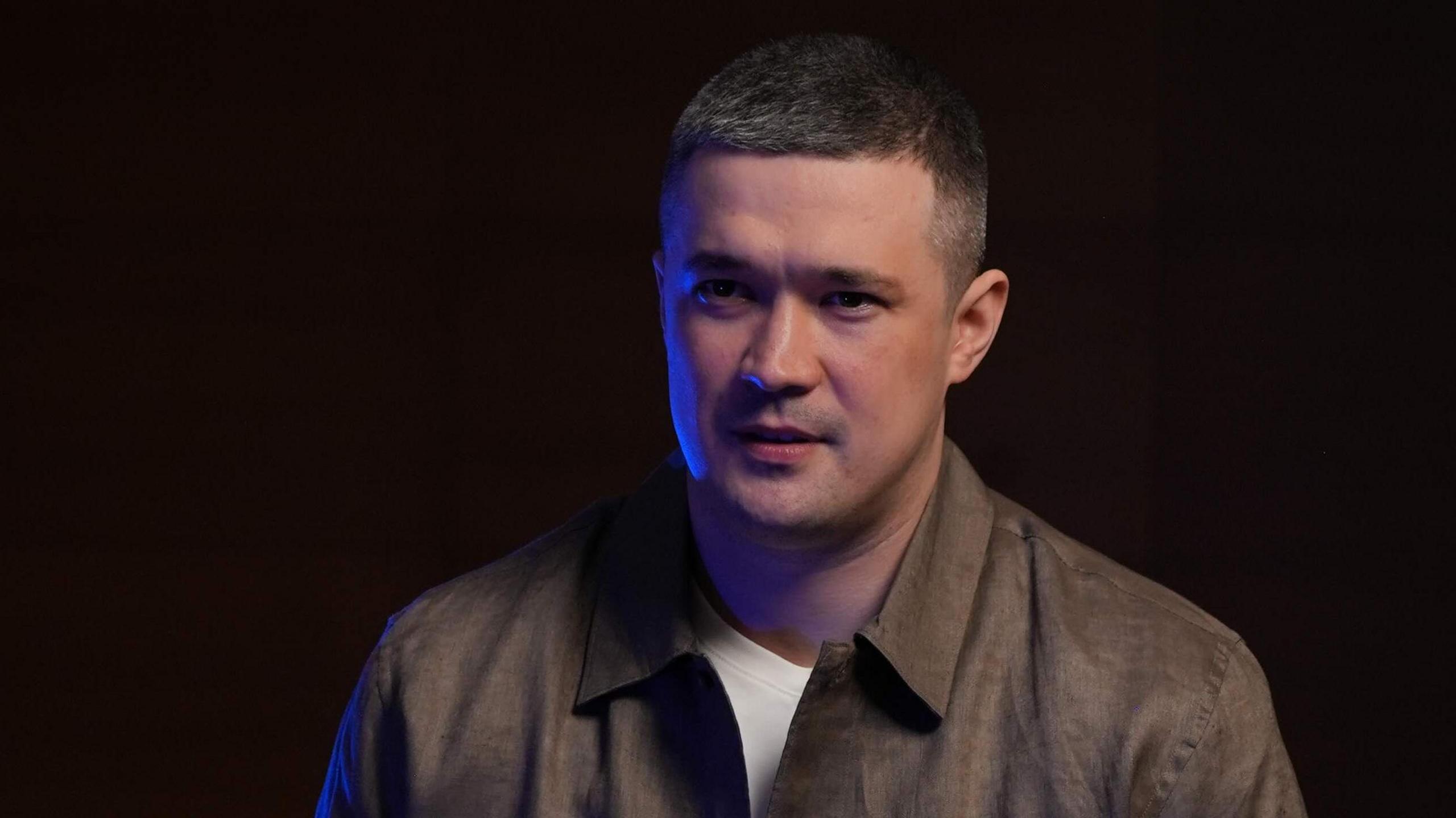 A man dressed in a white T-shirt and brown collared shirt in front of a black background sits talking to someone with their back to the camera.