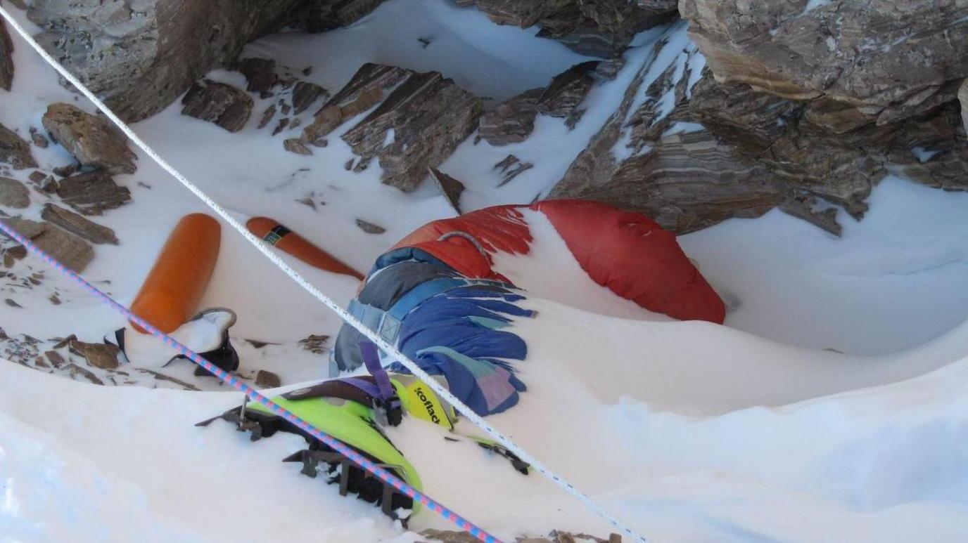 The dead body of a climber lying face down and partly covered by snow. His green boots can be seen on his feet.