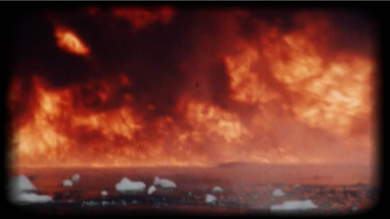 Archive-style footage shows an intense fire burning across a distant shoreline. Towering flames and smoke dominate the frame, while floating chunks of a white substance drift across the dark water in the foreground.