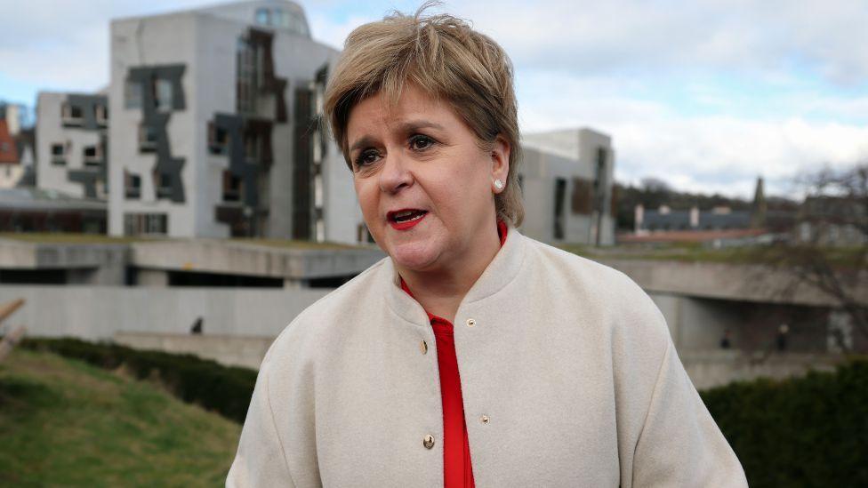 Nicola Sturgeon, who has short, fair hair, speaks outside the Scottish Parliament.  She is wearing a beige jacket with a red top underneath