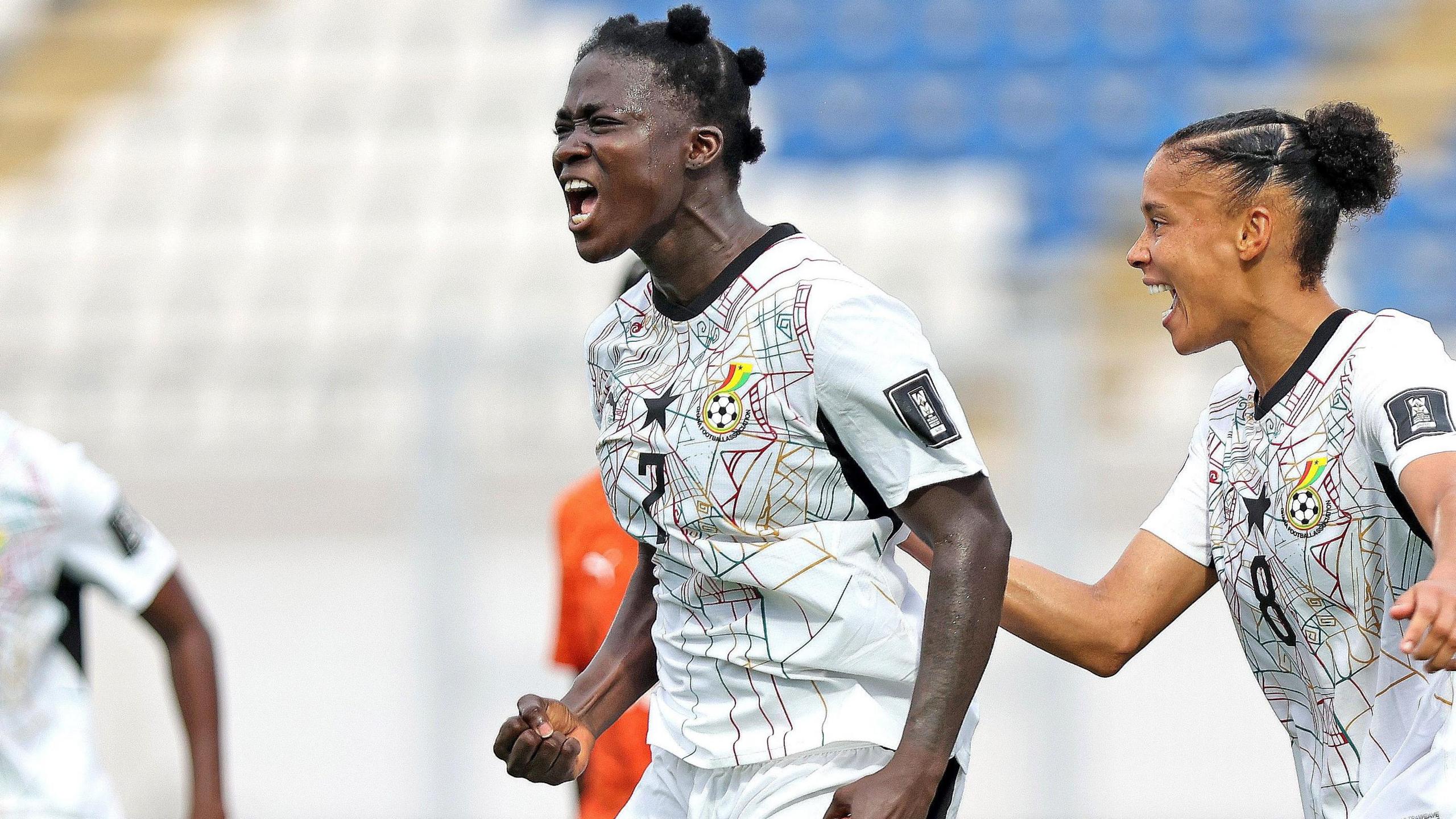 Princess Marfo clenches her right fist and screams in delight as she celebrates a goal for Ghana in the 2027 Women's World Cup play-off against Ivory Coast. Ghana team-mate Chantelle Boye-Hlorkah is seen behind Marfo joining in the celebrations. Both players are visible from waist up wearing white Ghana shirts with an elaborate multi-coloured spiderweb pattern on the front. Out of focus in the background is an empty stand full of blue and white seats