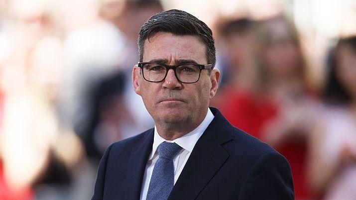 Andy Burnham wearing a dark navy suit jacket, white shirt and blue tie and dark-rimmed glasses - a crowd of people, not in focus, are behind him