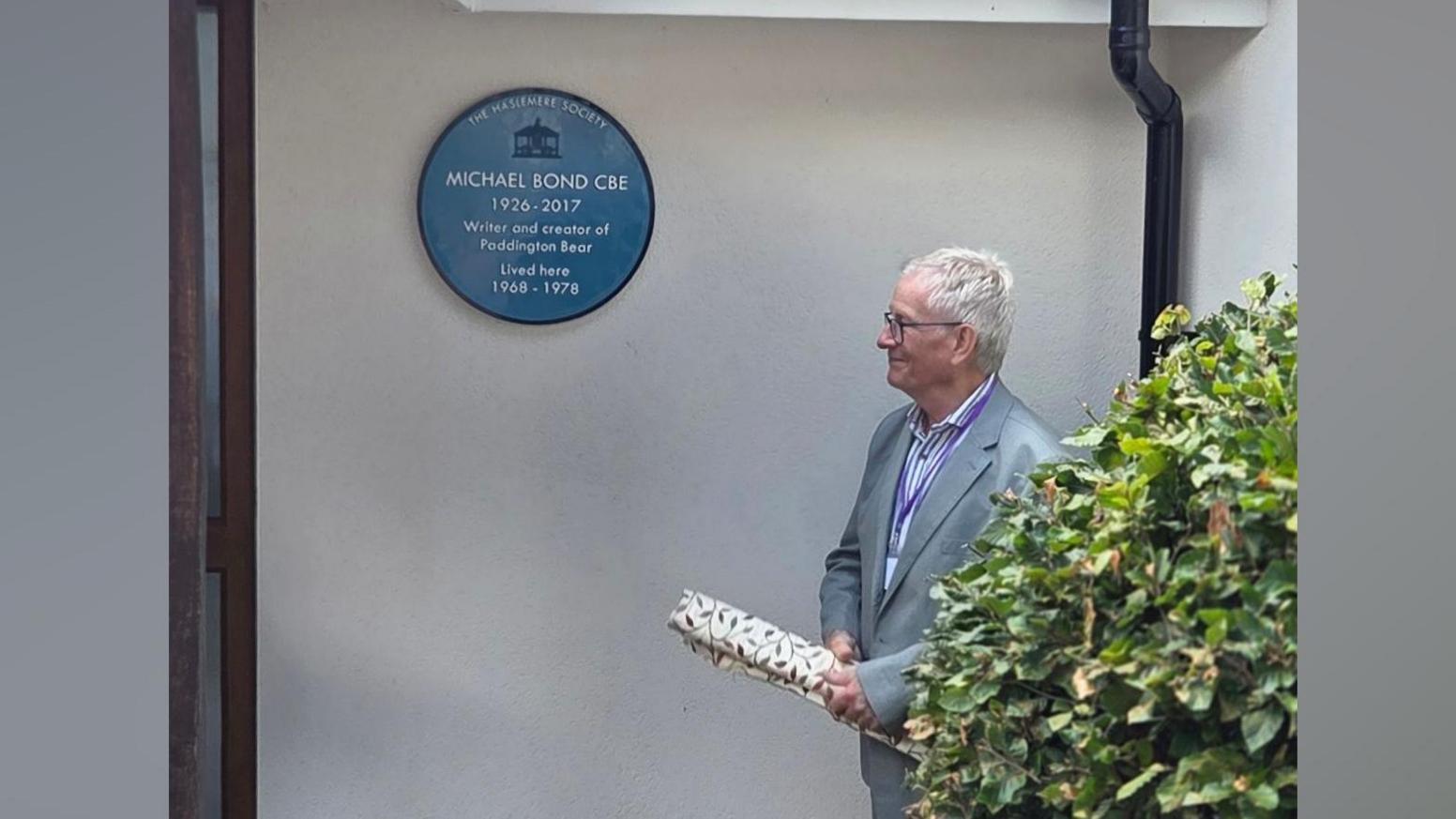 Chris Harrison, chair of the Haslemere Society, unveiling the blue plaque at the former home of Paddington Bear author, Michael Bond. Chris is in a grey suit and is holding the curtain used to cover the plaque. The blue plaque is visible in the image.