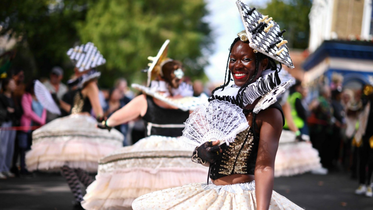 A woman in a pale coloured skirt and black and gold sequinned top and a headdress inspired by a chess board holds a white lace fan and smiles.