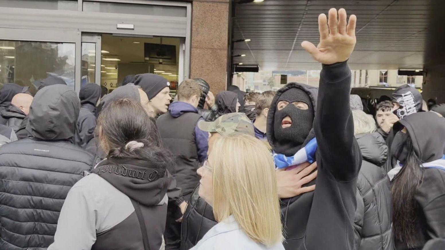 Crowd gathered outside a building entrance, with several people in dark hooded jackets and one individual raising an arm while holding a scarf around the neck, others recording with smartphones.