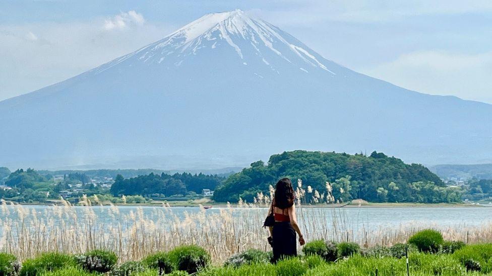 A photo taken from behind of a woman looking up at a snow-topped Mount Fuji, with a lake in front of it