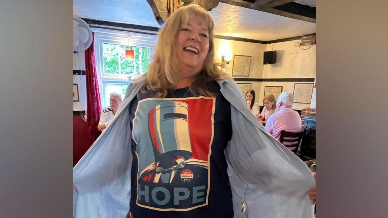 A woman proudly showing off her Count Binface HOPE T-shirt while standing in a pub. She is smiling and looking into the camera.