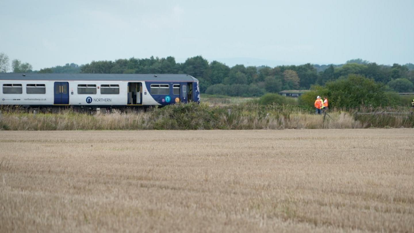 Image shows a train stopped on a railway line, with fields on either side. An official in a hi-vis jacket can be seen standing in front of it.