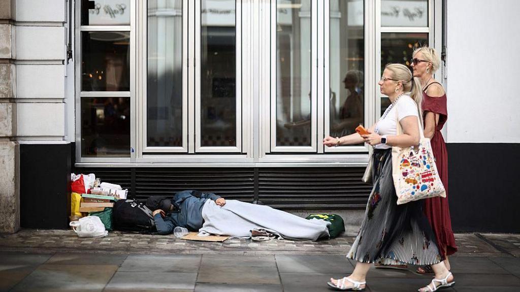 Two women walk past a person lying on the pavement in London, 28 July