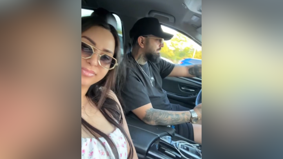 A woman and a man in a selfie-style video in the car. The man is driving and the woman is smiling into the camera. They are both wearing sunglasses.