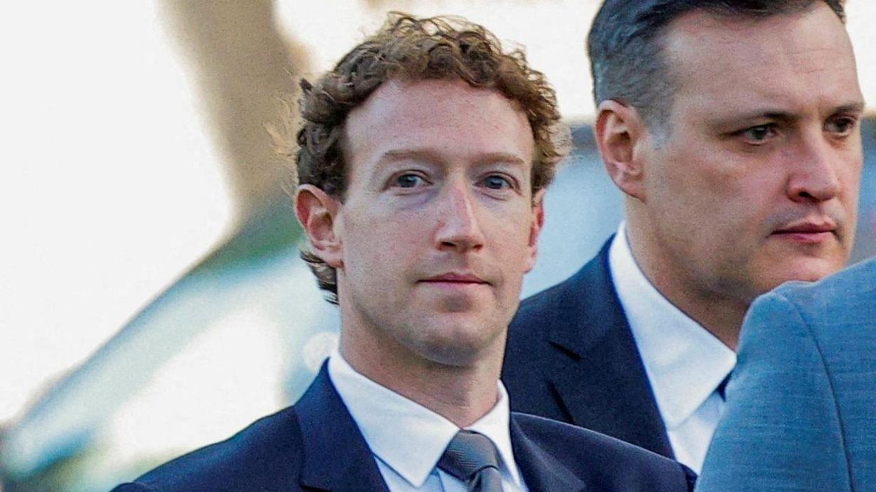 Mark Zuckerberg wearing a suit, looks towards the camera with a blank expression. He is flanked by staff also in suits.
