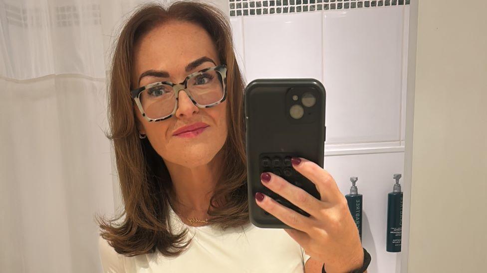 A woman with straight brown hair, a white t-shirt, patterned glasses and pink lipstick takes a selfie in the mirror with a phone