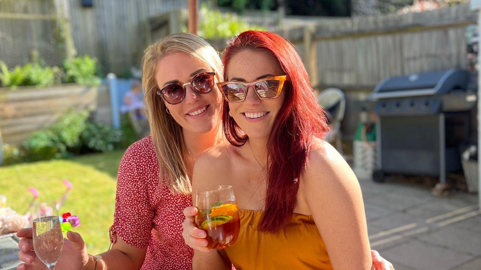 Friends Sarah (left) and Leanne (right) wearing sunglasses, smiling and holding drinks as they sit in a garden.