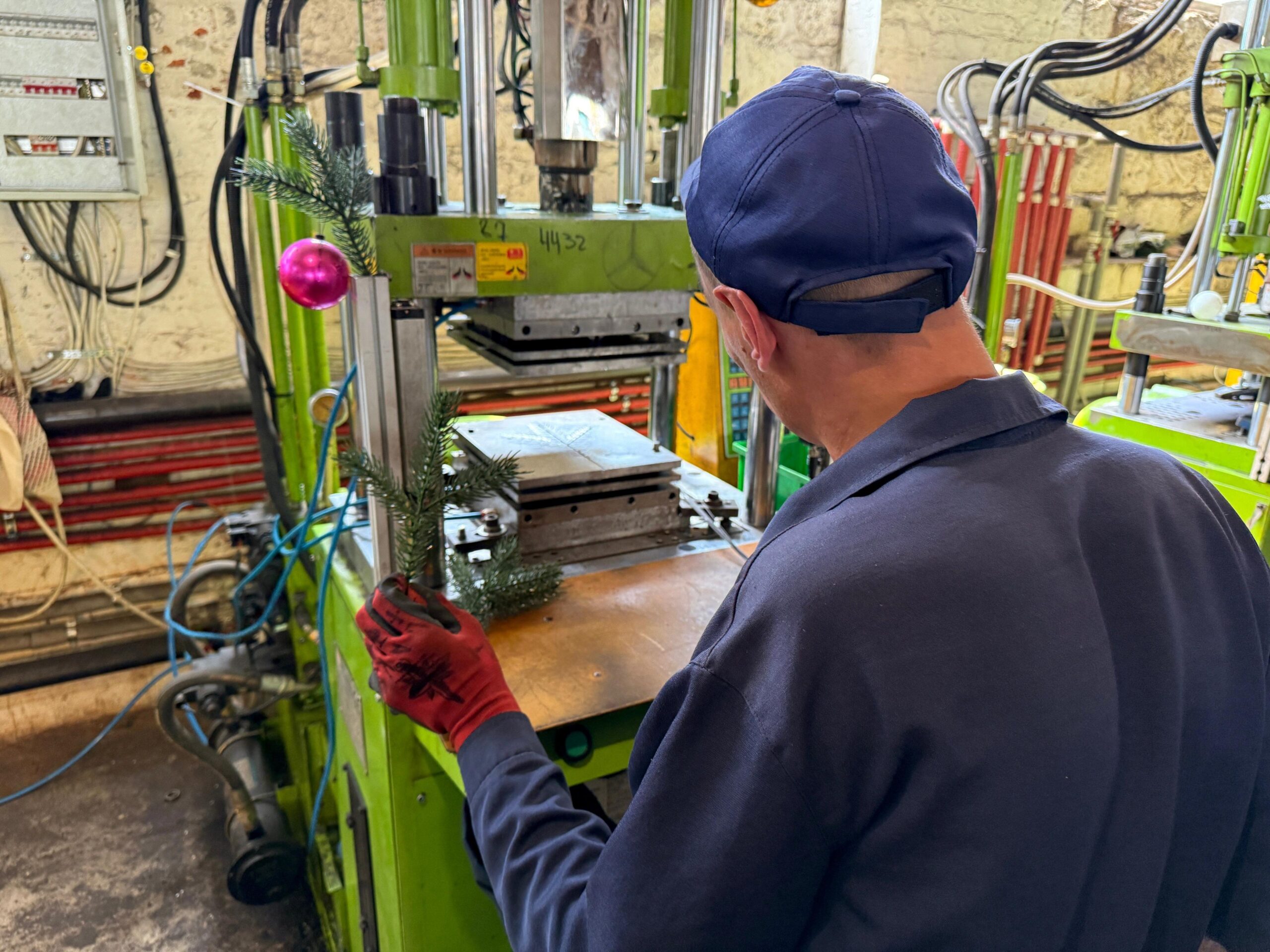 The back of a man using machinery to make Christmas decorations. There is the branch of a fake Christmas tree in his hand a pink bauble handing on the machine.