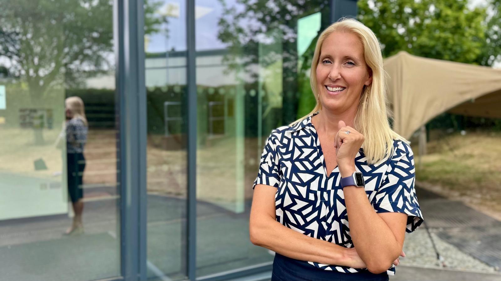 Vicky Orsmond is seen standing outside a modern glass-fronted building, wearing a patterned short-sleeved shirt and a smartwatch. A reflection is visible in the window behind, with trees, a canopy structure and landscaped grounds in the background.