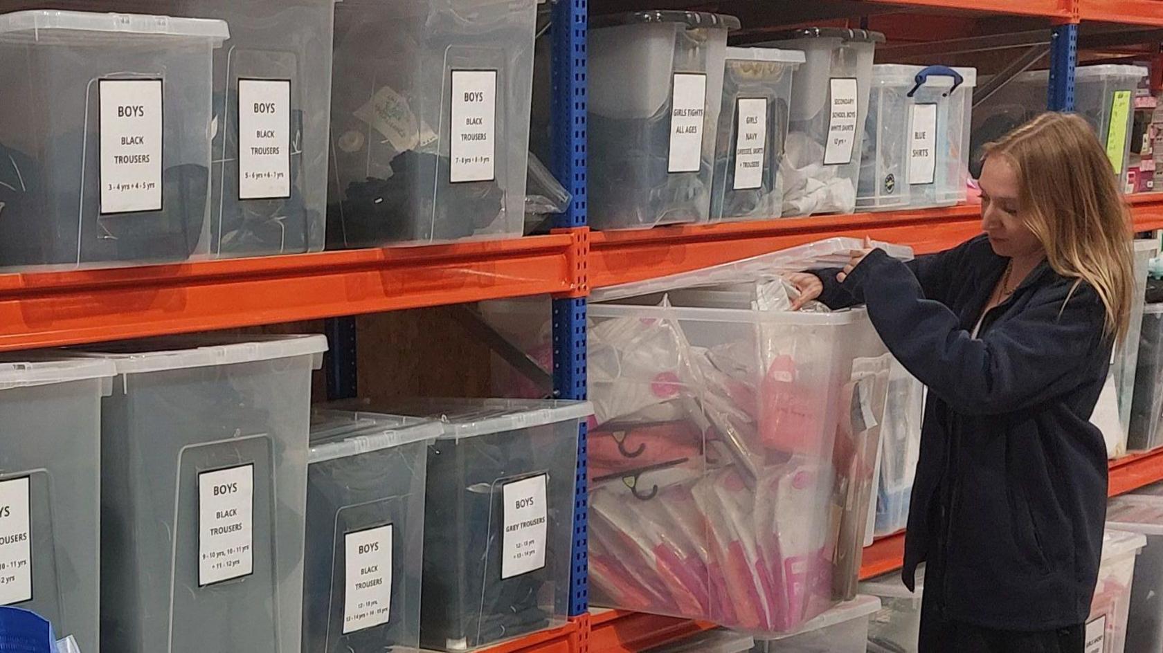 A volunteer stands next to boxes of boys' black and grey school trousers.