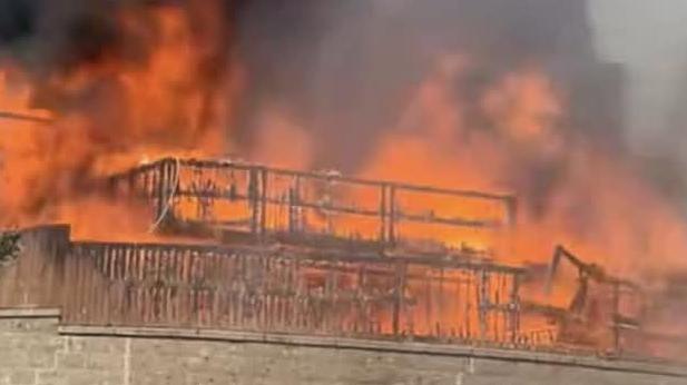A burning fence and the frame of a house is all that is visible in this picture from  below a wall of orange flames. Black smoke is coming up from a bungalow above a grey brick wall.