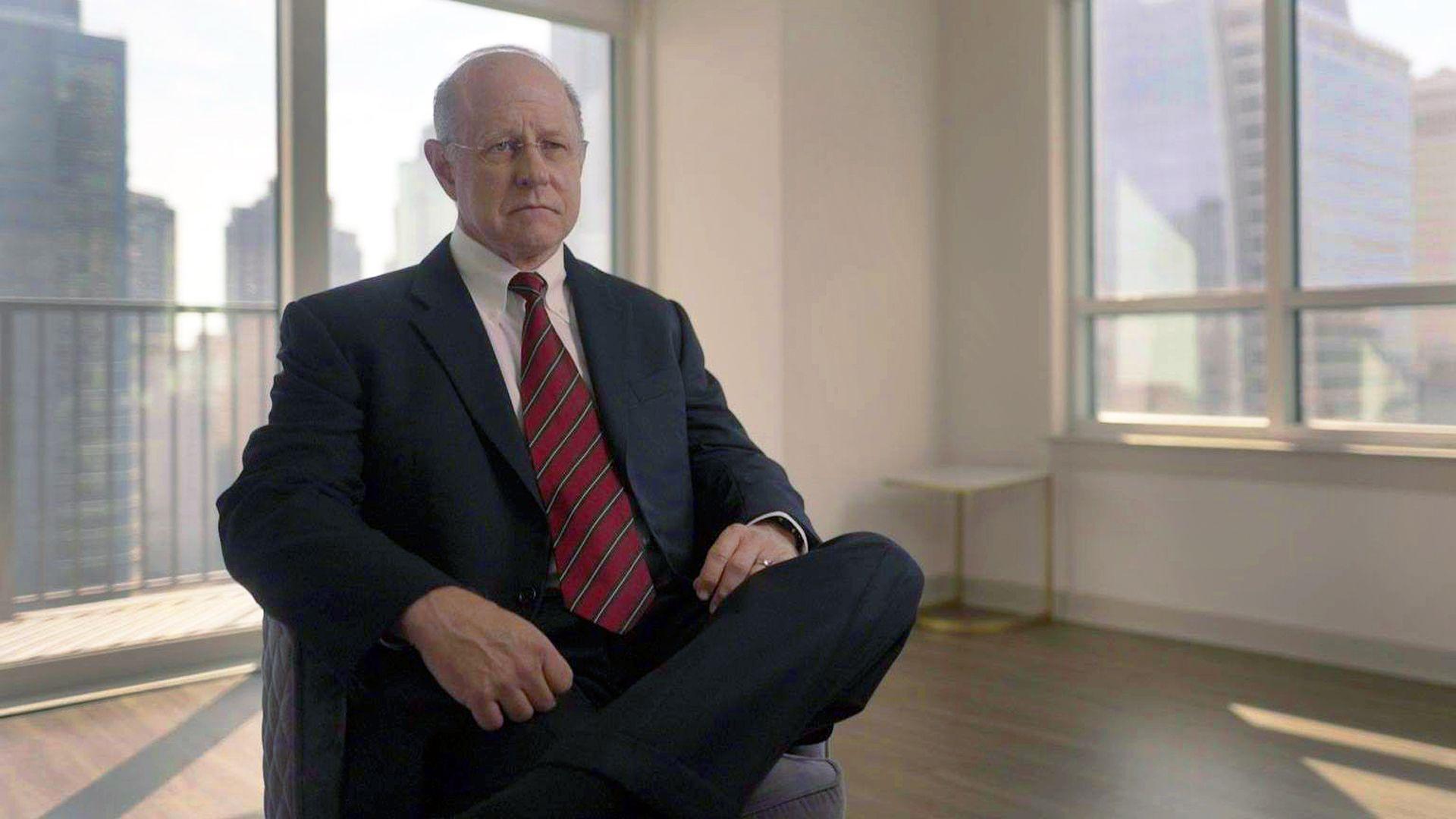 Richard Lambert, who has now retired from the FBI, is interviewed in an office by the BBC. He wears a suit with a white shirt and red tie and has his legs crossed.