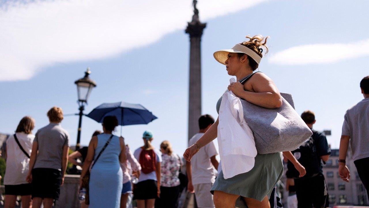 A woman with a sunhat and visor on walks through Trafalgar Square with other people holding umbrellas in the background to shield them from the sun. With Nelson's Column in the background