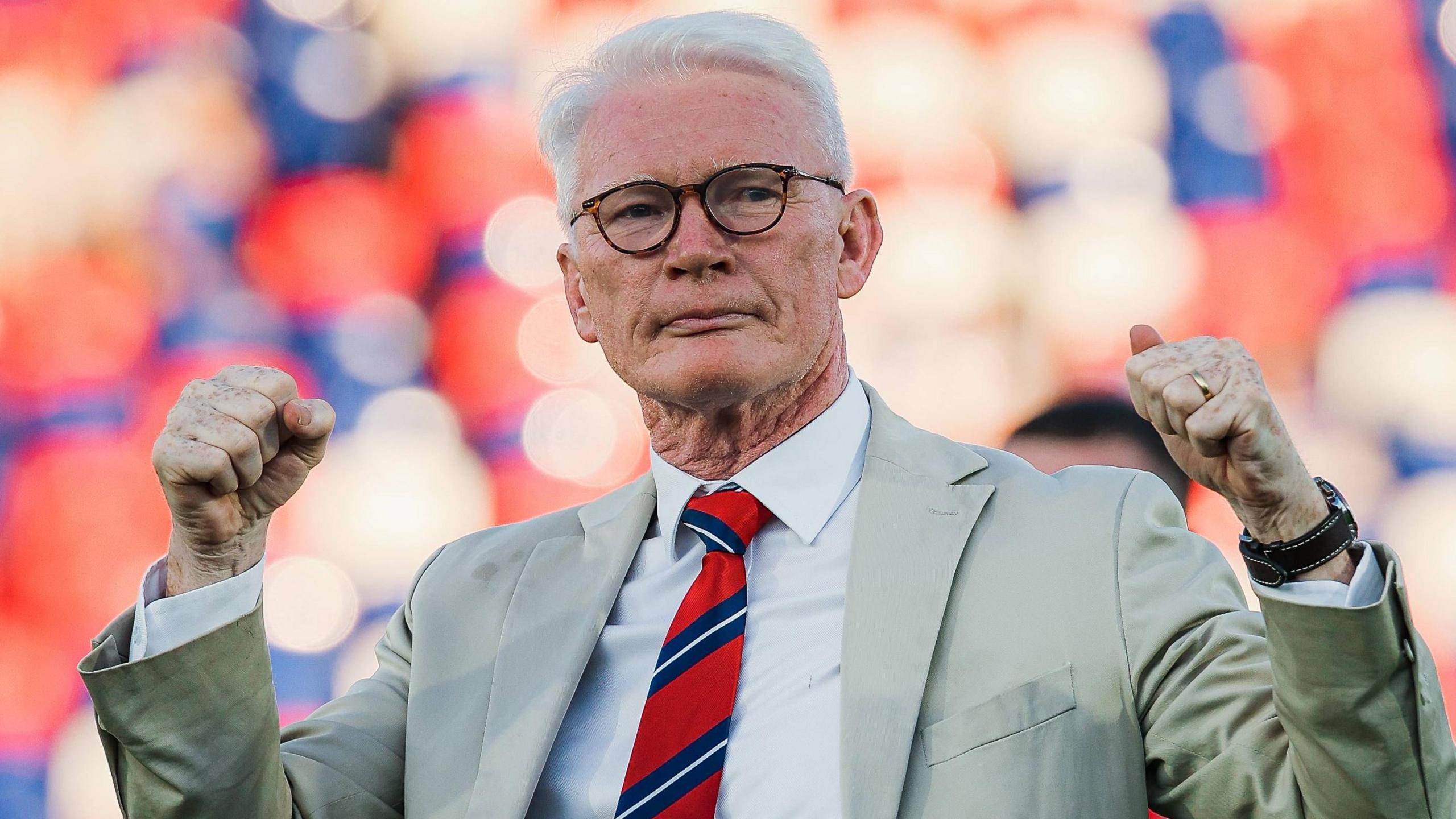 Eamonn McManus, wearing a beige suit jacket, light coloured shirt and red and blue tie, pumps both his fists in the air to celebrate after a St Helens game