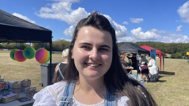 Chloe Fogg pictured at the event with long brown hair and wearing blue denim dungarees and a white top. Behind her are stalls and people gathered around them.