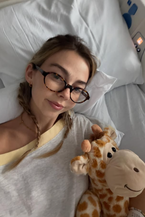 Towle, wearing glasses and a t-shirt, laying down on a pillow in a hospital bed with a giraffe stuffed animal