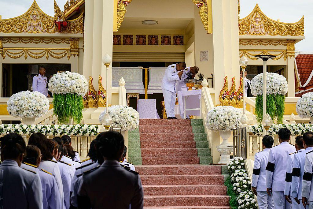 Mourners gather by a crematorium decorated with white floral arrangements as uniformed attendees stand on either side of a staircase. At the top of the steps, two officials stand beside white coffins.